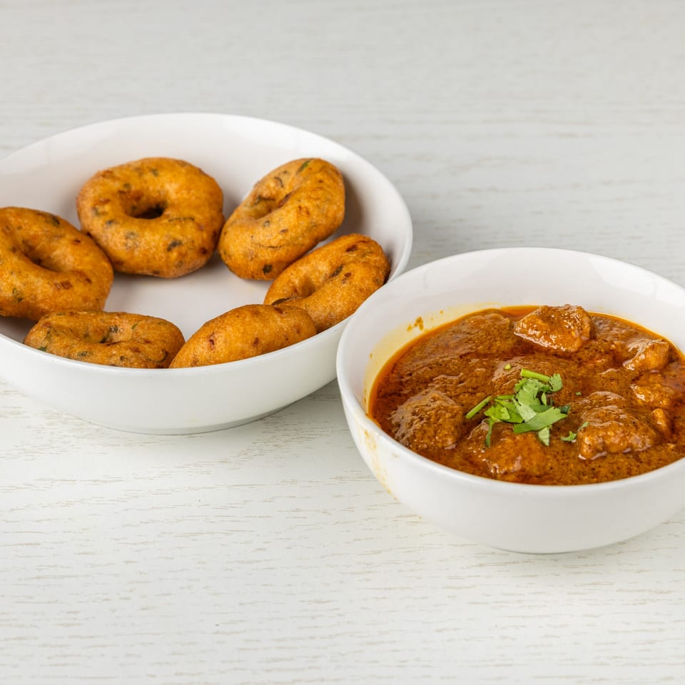 A bowl of chicken curry with cilantro and a plate of golden-brown medu vada on a white table.