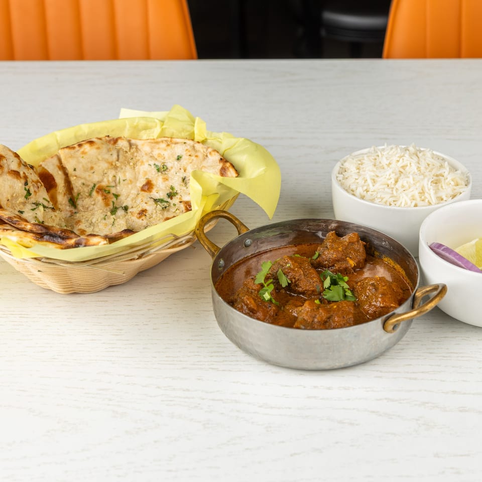 Indian lamb curry in a metal bowl, with naan bread in a basket and white rice.